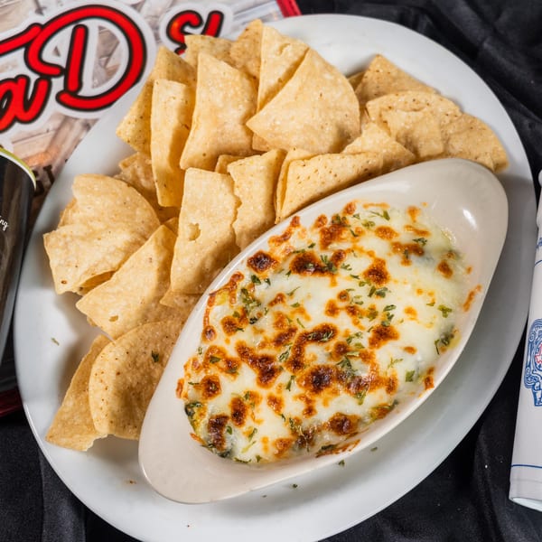 A white plate holds a boat-shaped dish of cheesy dip with tortilla chips, a drink, and seasoning.