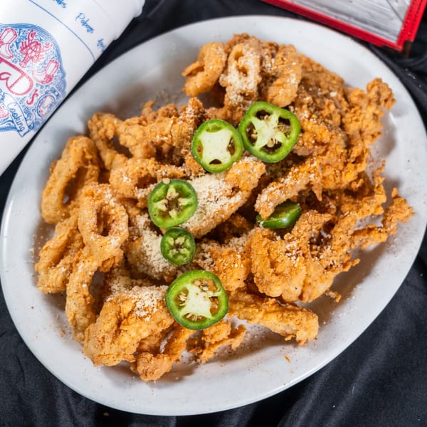 A white plate of golden fried alligator with parmesan cheese and jalapeño slices, next to a Taj's cup.