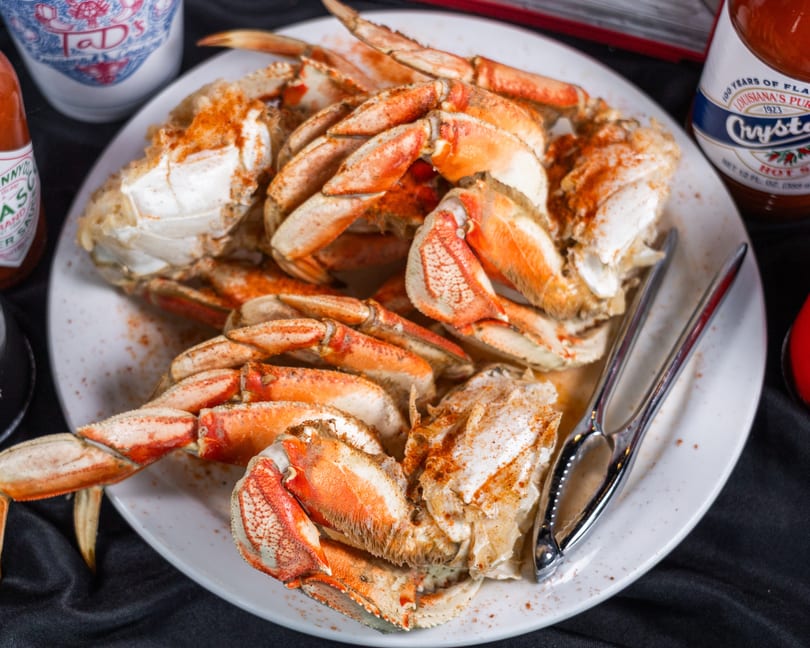 A plate of cooked crab legs and claws seasoned with spices, with hot sauce bottles nearby.
