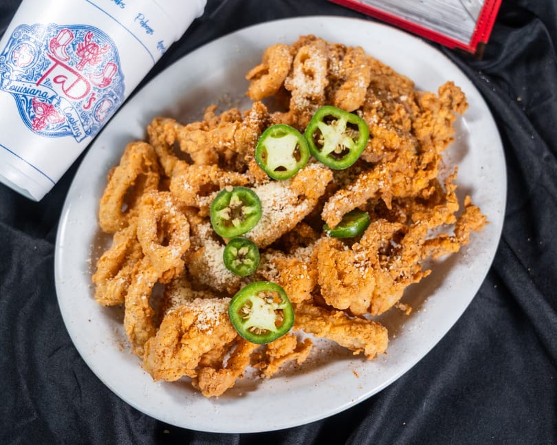 Fried alligator with parmesan cheese and jalapeño slices on a white plate next to a Taj's cup.