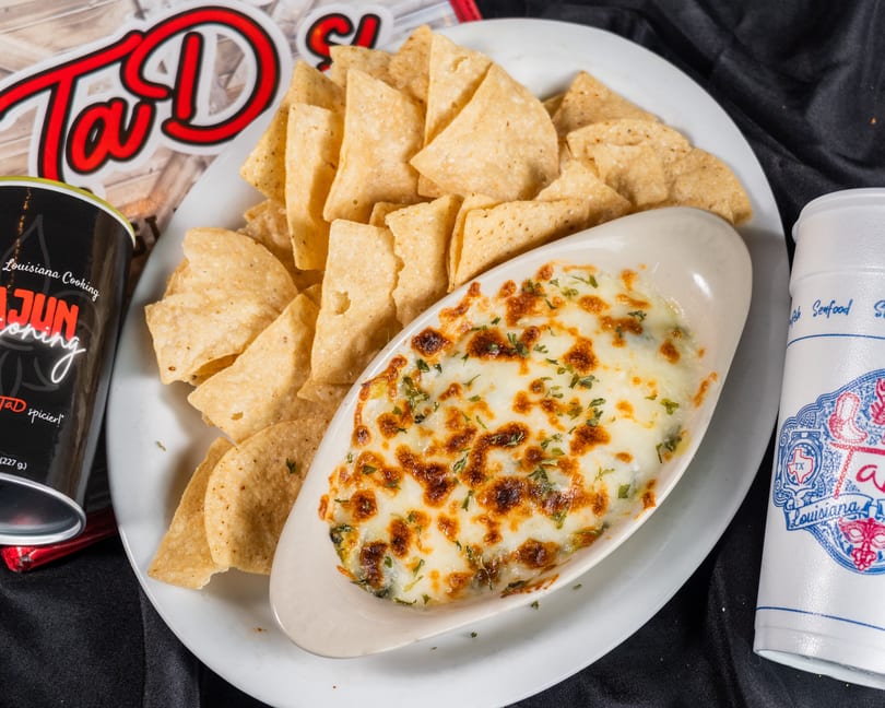A white platter holds a boat-shaped dish of baked spinach dip with tortilla chips, a drink, and seasoning.