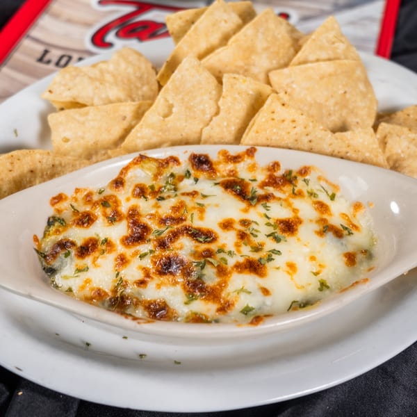 A white oval dish of melted cheese dip with herbs, surrounded by tortilla chips on a white plate.