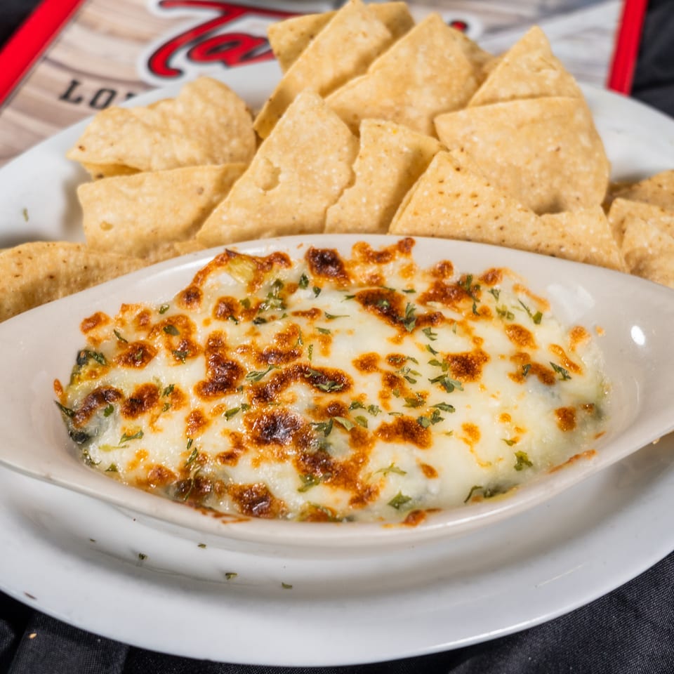 A white oval dish of melted cheese dip with herbs, surrounded by tortilla chips on a white plate.