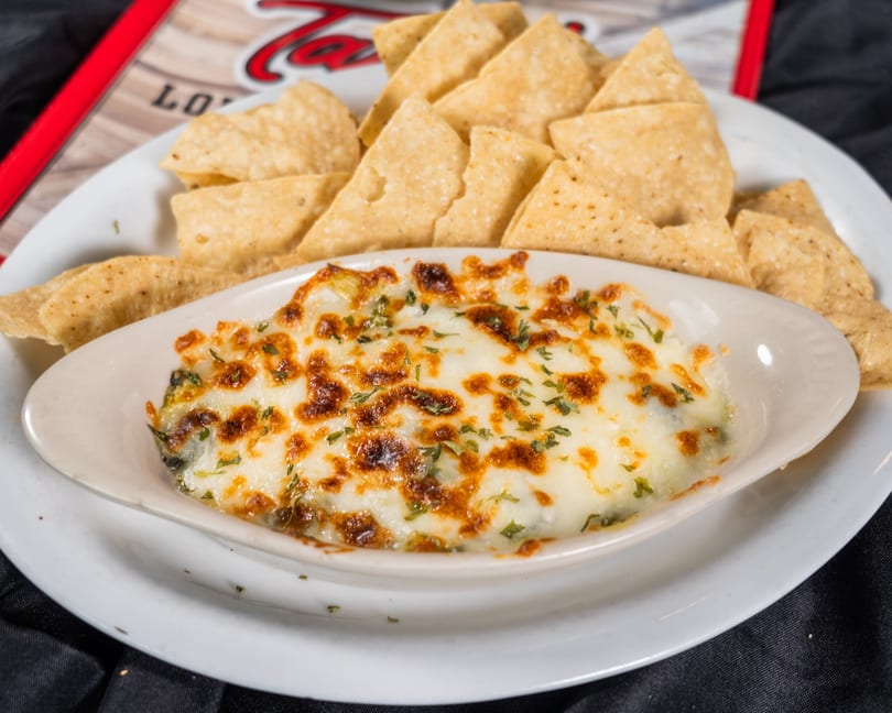 A white oval dish of melted cheese dip with herbs, surrounded by tortilla chips on a white plate.