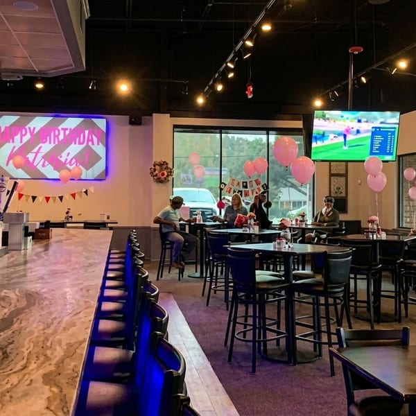 Restaurant interior with a long bar, high-top tables, and pink balloons for a birthday celebration.