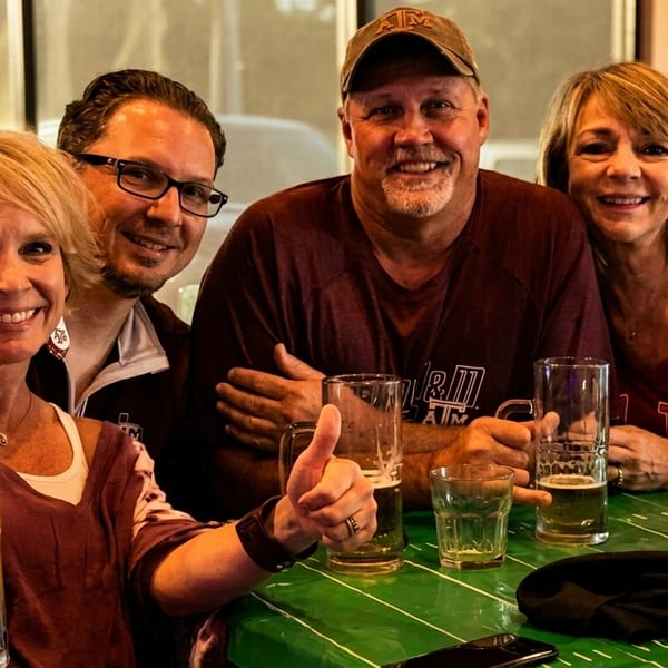 Four smiling adults, two men and two women, enjoying beers at a table with a green tablecloth.