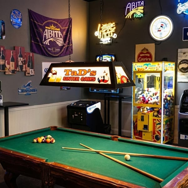 A game room with a pool table, arcade games, claw machine, and neon bar signs.