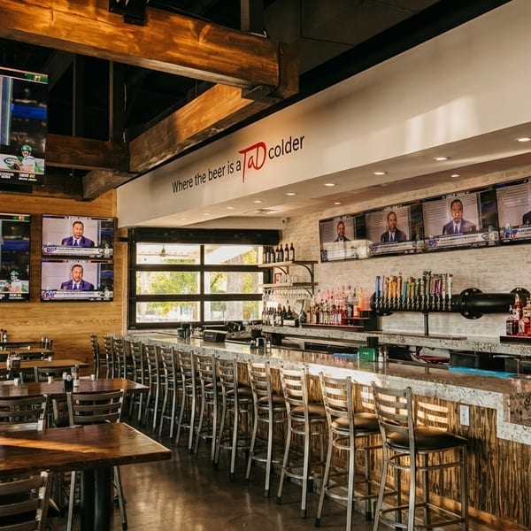 A sports bar interior with multiple TVs showing sports, a long bar with stools, and dining tables.